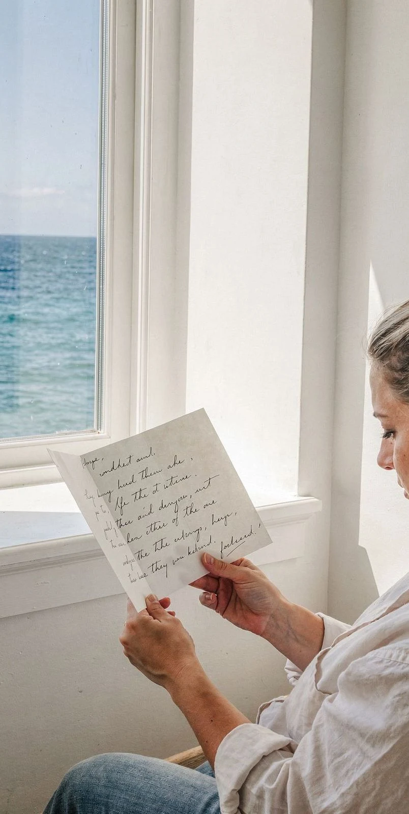 Una mujer lee junto a una ventana abierta al mar.